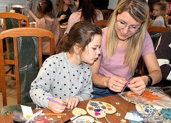 Dziewczynka z mamą. Mama przywiązuje sznurek. Na stole serwetki, nożyczki. pisaki, celofan i wstążki