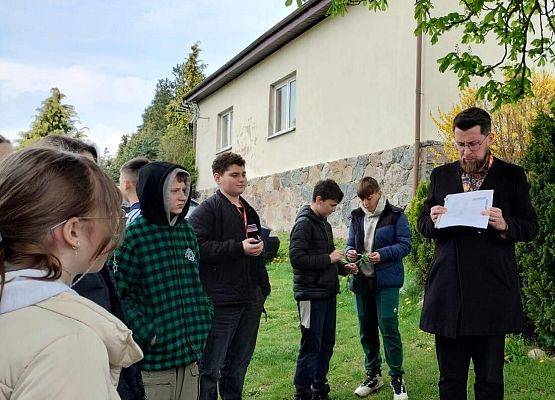 Po prawej edukator z tabelą w dłoniach, ku niemu skierowana grupa uczniów stojąca z lewej, w tle budynek szkolny