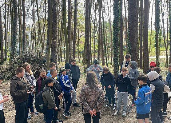 Zwarta grupa dzieci z instruktorem w parku, zaraz za nimi wysokie drzewa.