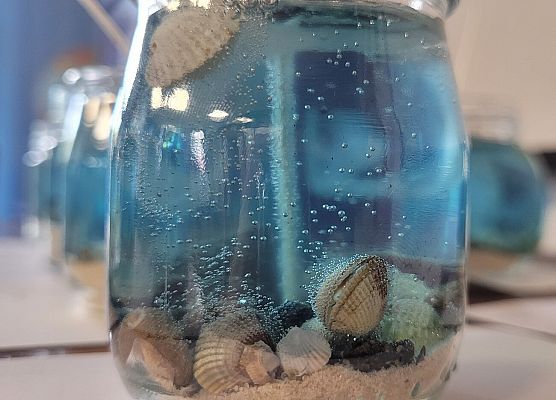 Świeczka żelowa imitująca dno morskie. Na dnie piasek, muszelki małży, kamyki zalanie niebieski żelem.