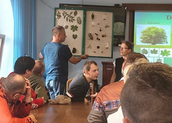 Grupa na sali edukacyjnej. W tle tablica z liśćmi i nasionami różnych gatunków rzzew
