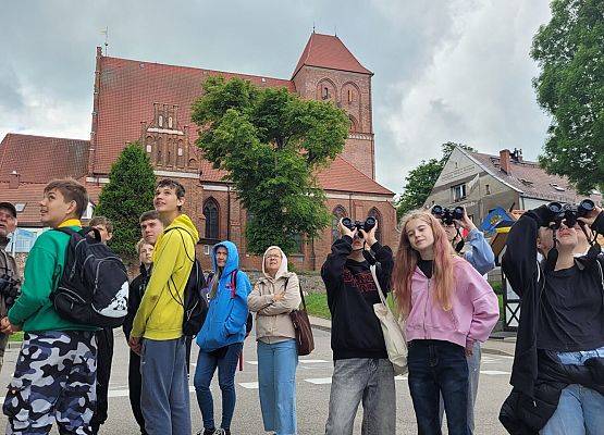 Uczniowie -ok 10 os. obserwują ptaki za pomocą lornetek. w tle kośiół farny w Pucku