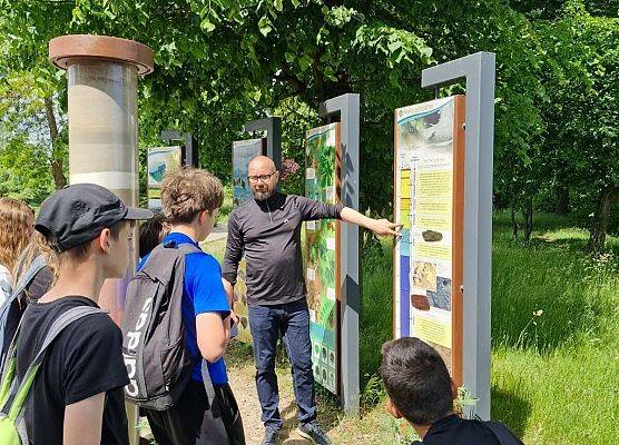 Grupa w terenie przy ekspozycji geologicznej edukacyjnej, W tle drzewa.
