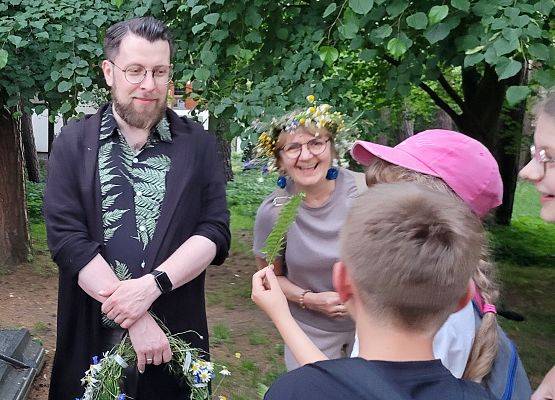 W tle drzewa. Dziecko trzyma gałązkę paprotki. Uśmiechnięta pani z wiankiem na glowie. Pan trzyma w rękach wianek